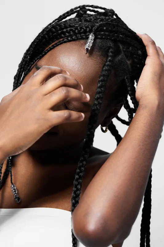 A young Black woman with braided hair expresses emotions while touching her face against a clean, minimalistic background emphasizing her beauty and style