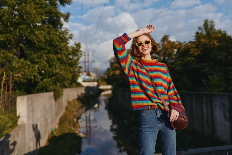 Young woman in a colorful striped sweater and sunglasses smiles on an urban bridge by a canal, mid-20s model in jeans and casual outfit with a round crossbody bag, raising hand to shade eyes.
