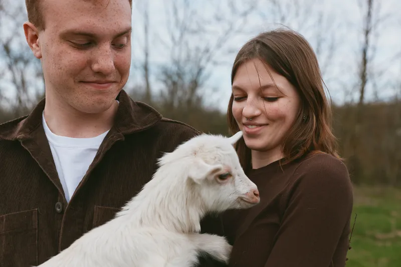 Young couple holds a white baby goat outdoors during autumn while sharing a tender moment in casual clothing and natural light in a countryside lifestyle setting.