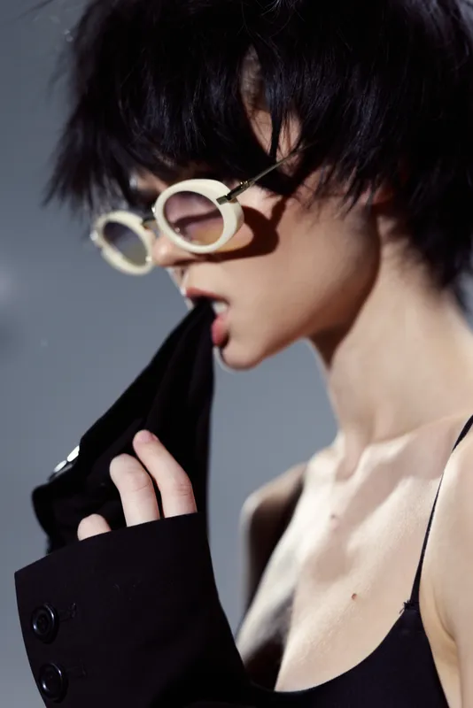 Stylish young woman wearing round glasses biting a black jacket sleeve, with short dark hair and a soft, moody background.