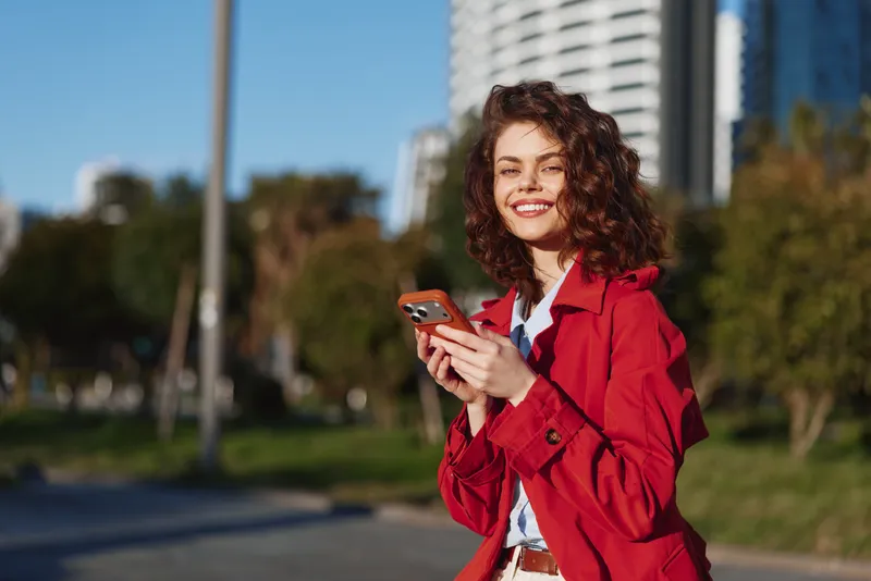 Young woman in a red coat smiles while checking her phone in a sunlit city park with modern buildings behind, a soft breeze ruffling her hair and green grass around