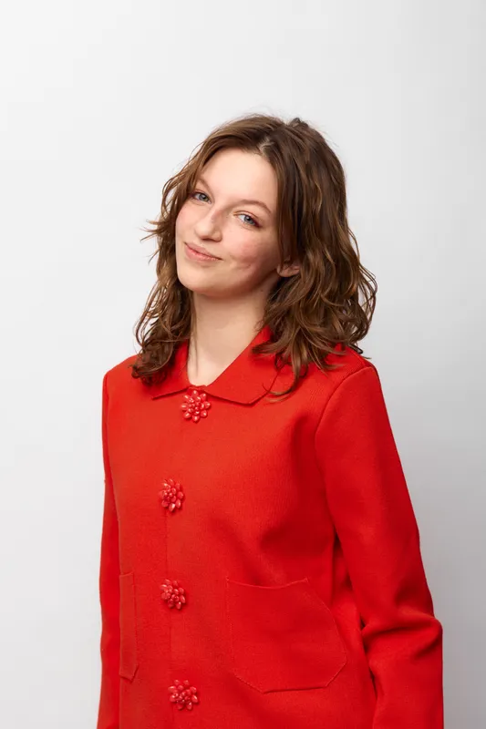 Young woman with curly brown hair wearing a red sweater smiles softly against a white background. Casual portrait with natural light and relaxed confident expression.