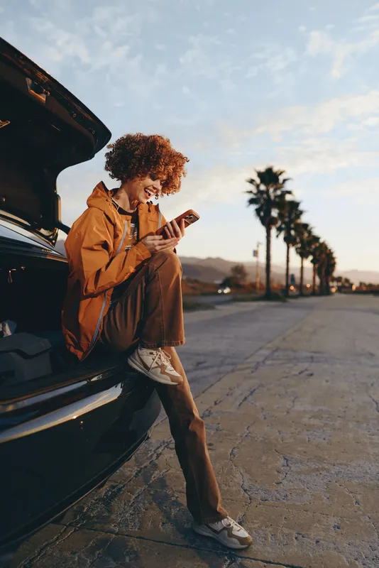Woman lifestyle travel outdoors, roadside car, sunset pavement, she is seated by the trunk, looking at phone while packing suitcase, casual jacket, sneakers, relaxed mood, evening