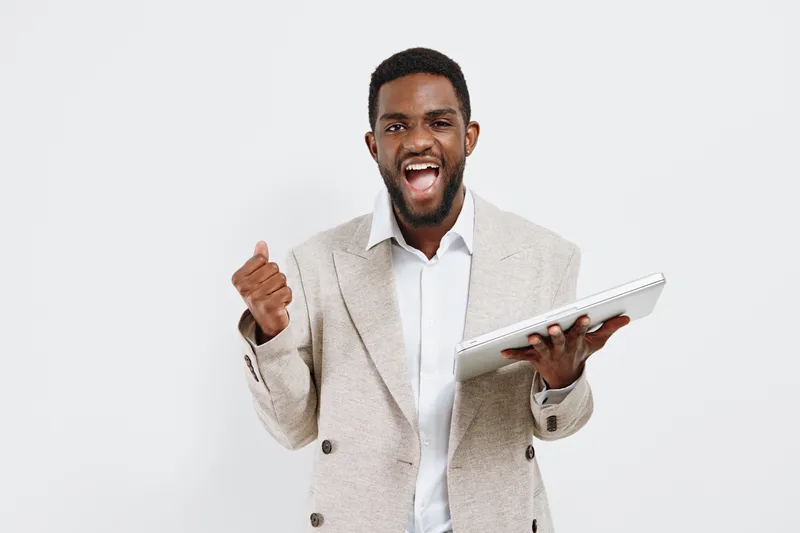 Joyful smiling man with dark skin wearing beige blazer and white shirt holding tablet computer on plain white background. People lifestyle concept, celebration, success, achievement, happiness, positi