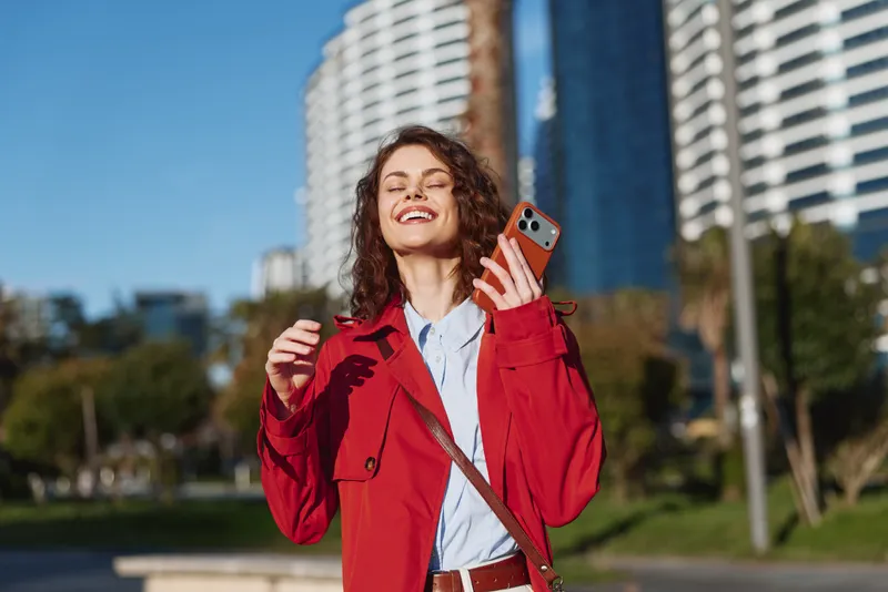 A cheerful woman in a red coat takes a selfie in an urban park, bright skyline behind. A candid portrait conveying confidence, joy, and modern city life.