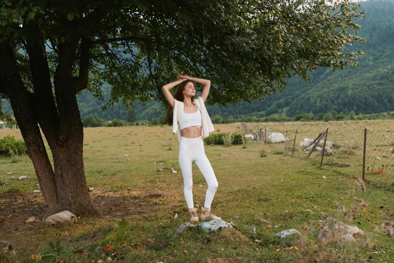 Young woman in white activewear stands on a rock under a tree in a rural field enjoying nature and fresh air with a relaxed and confident expression.