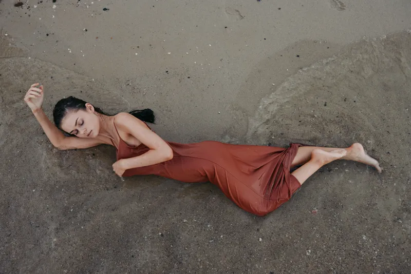 A graceful woman lies on damp sand wearing a flowing red dress, creating a dramatic silhouette and a serene, sunlit mood on a quiet beach.