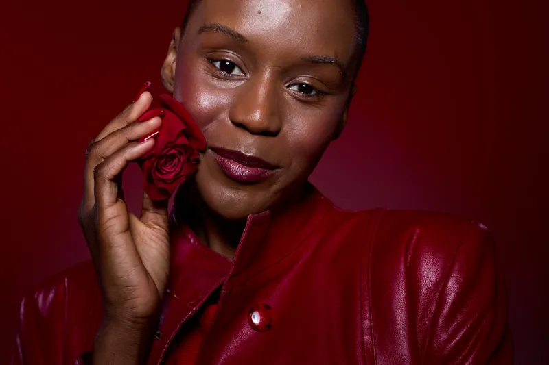 Close-up portrait of a dark skinned African woman holding a red rose near her face. She wears a red leather jacket against a deep red background. Concept of beauty, elegance, and confidence.