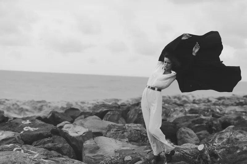 Woman on rocky shore, cape billowing in wind, embracing the open sea in a dramatic fashion moment, capturing movement and freedom by the coast.