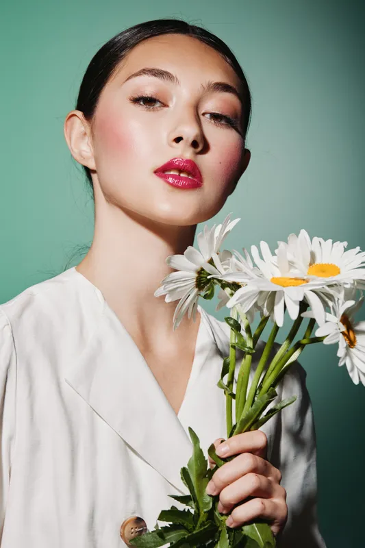 Fashion model with vibrant makeup holding daisies looks confidently at the camera wearing white outfit. Studio shot on green background in artistic beauty concept.
