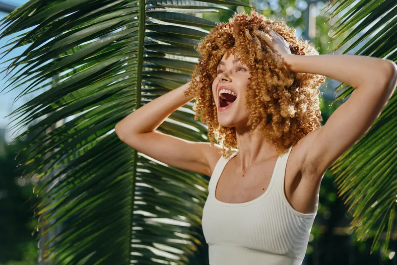 Joyful woman with curly hair enjoying tropical nature, smiling brightly under green palm leaves in warm sunlight, summer casual style vibrant energy.