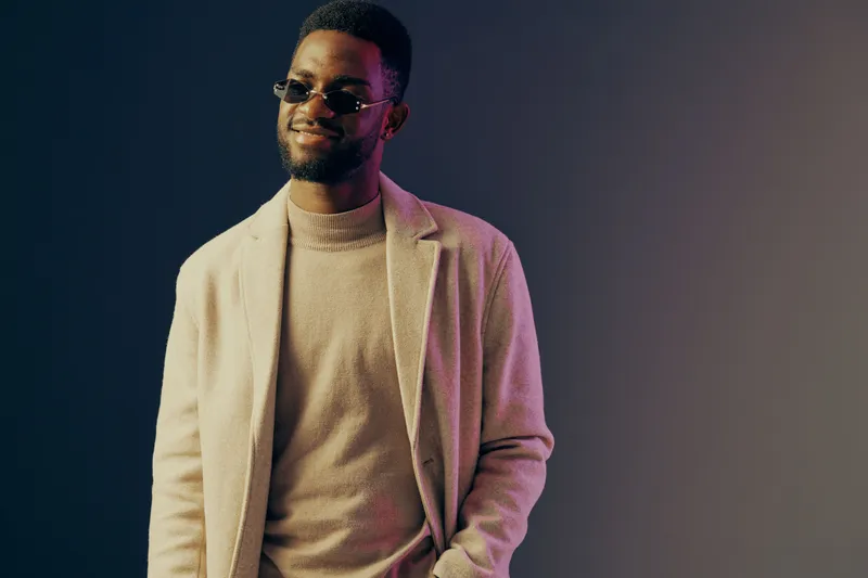 Stylish African man in sunglasses wearing a beige sweater and blazer, smiling against a colorful gradient background, embodying modern fashion and confidence