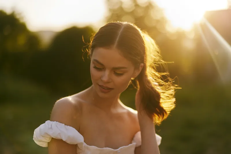 Young woman with long hair enjoying a sunny day outdoors, wearing a white off shoulder dress, radiating a serene and thoughtful emotion