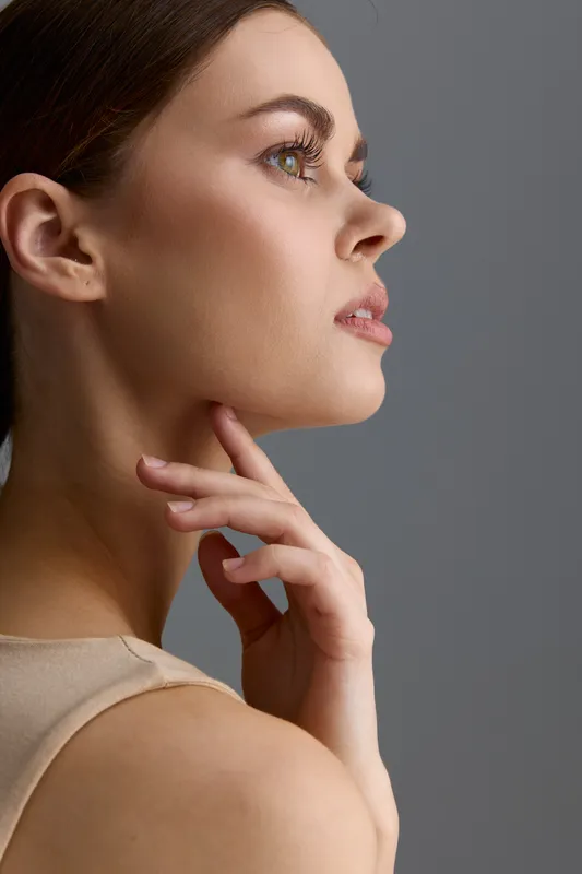 Beauty woman with clear skin and natural makeup posing in profile against gray background, showcasing soft expression and delicate hand gesture in minimalist style photography.
