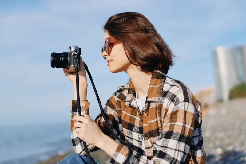 Photographer in plaid shirt by the seaside with a camera, capturing coastal scenery and candid moments. Outdoor portrait with sunny weather, sunglasses, and a calm shoreline.