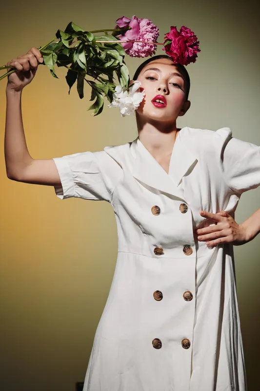 Fashion model poses with pink peonies against muted background wearing white dress with oversized buttons. Artistic beauty and creative fashion theme emphasizing elegance and floral style.