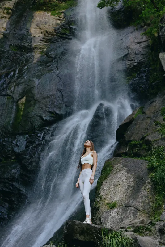 Woman in white sportswear standing near a tall waterfall surrounded by mossy rocks and lush green foliage in a serene natural environment.