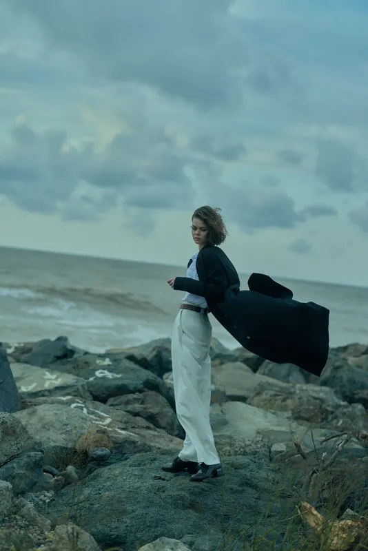 Woman stands on a rocky coast by the sea, wearing a dark coat and light trousers, wind catching the fabric, moody clouds above, creating a dramatic coastal atmosphere
