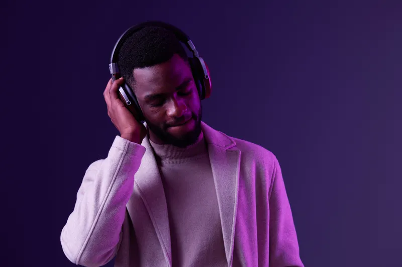 Stylish young Black man in headphones enjoying music against a purple background, showcasing modern fashion and relaxation