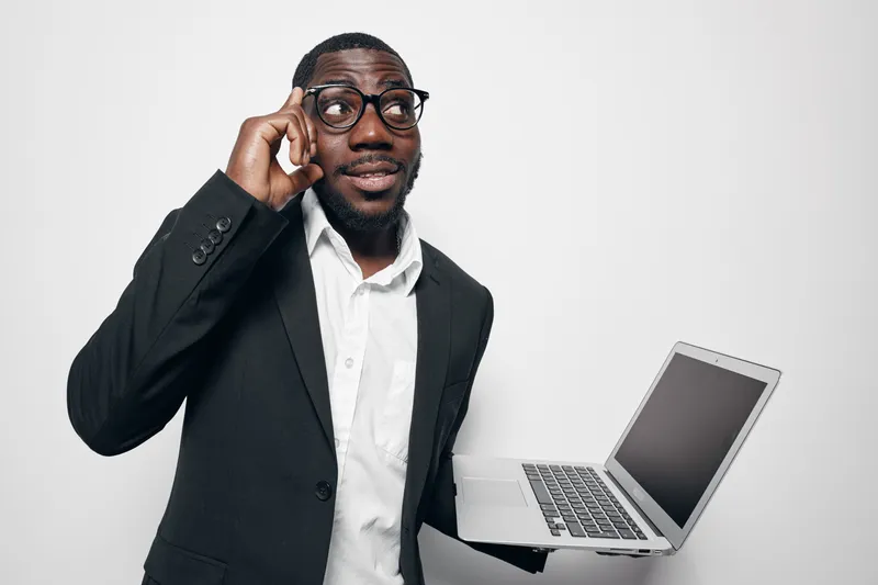 African American man with dark skin wearing glasses and black suit holding laptop and pointing to head with thoughtful expression on white background.