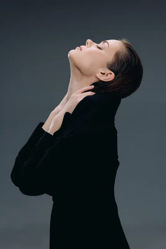 Elegant woman in black outfit with closed eyes and graceful pose on dark gray background.