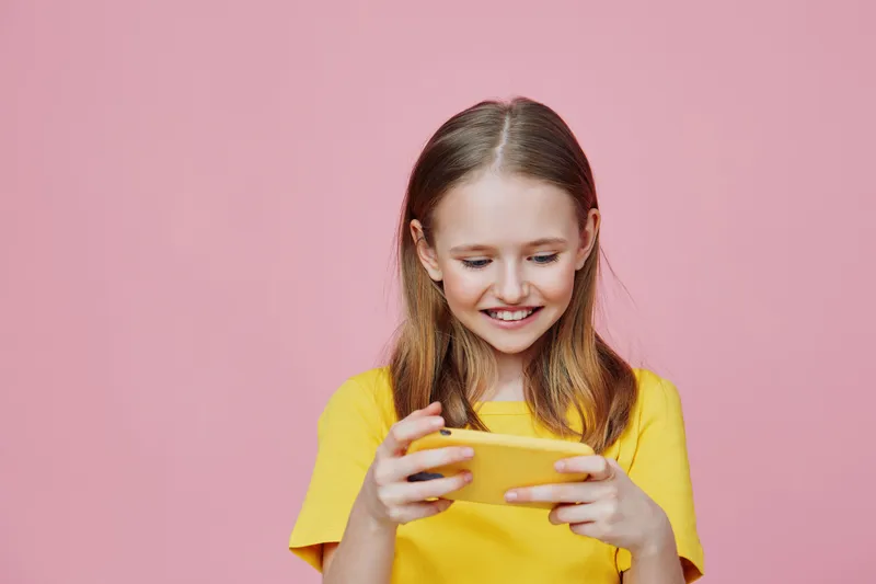 Happy girl playing on smartphone against pink background, wearing a bright yellow t shirt, showcasing joy and engagement with technology