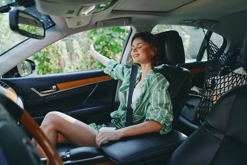 Young woman relaxing in a car, smiling with her seatbelt fastened, leaning toward the window as sunlight streams through the cabin.