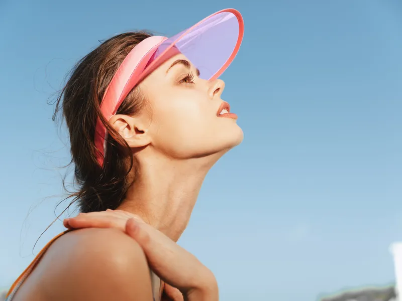 Portrait of a woman wearing a pink translucent visor, gazing upward against a clear blue sky, sunlight highlighting her relaxed features and casual summer mood.