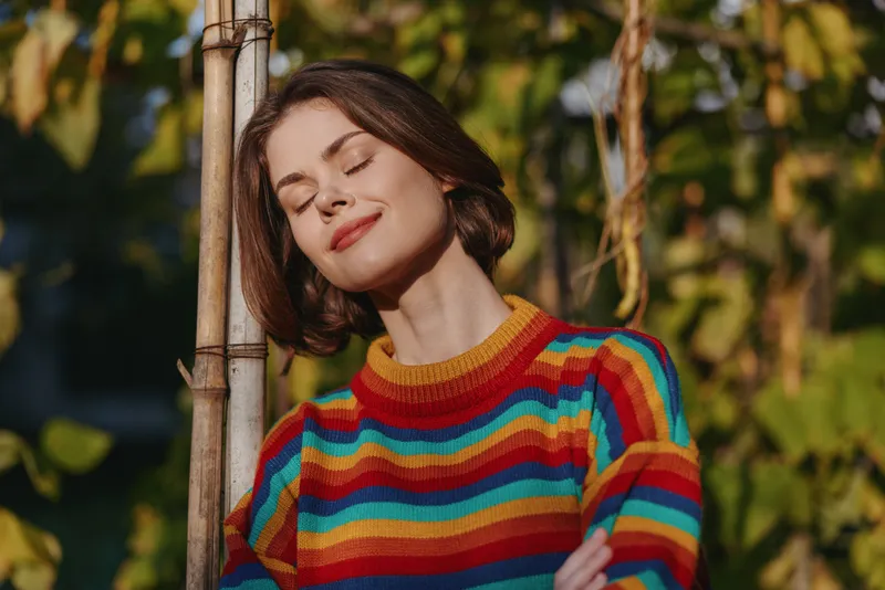 woman portrait smile colorful sweater closed eyes outdoor, young woman leaning on a pole with content expression and short hair, warm sunlight, relaxed mood and nature lifestyle scene