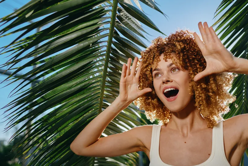 Young woman with curly hair expressing excitement outdoors under bright blue sky with tropical palm leaves creating vibrant summer atmosphere.