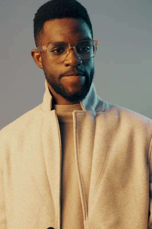Stylish young man in a light coat with glasses, showcasing confidence against a blue and beige background, ideal for fashion and lifestyle photography