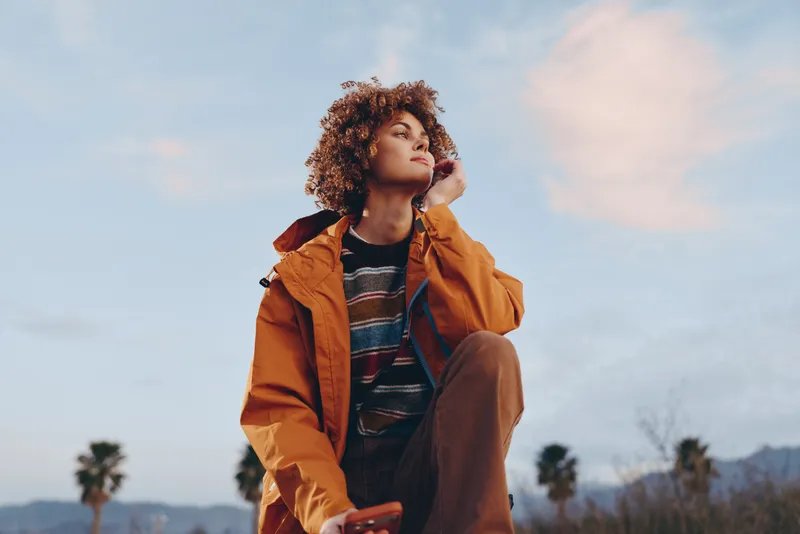 Woman lifestyle outdoor portrait, curly hair, orange jacket, adjusting collar, gazing upward, calm thoughtful mood under pastel sky, distant palm trees, natural soft daylight