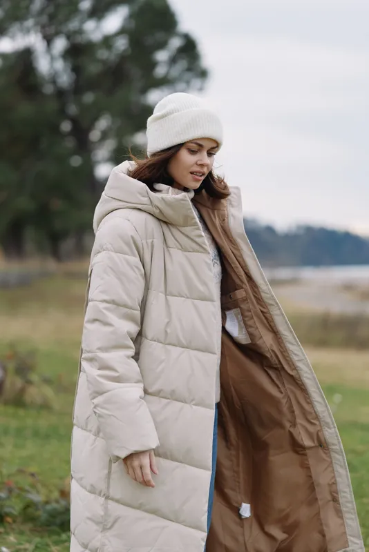 Woman wearing a light beige puffer coat standing outdoors in a grassy landscape, bundled in a knit hat, with the coat open to reveal a warm interior and casual attire for a ready look.