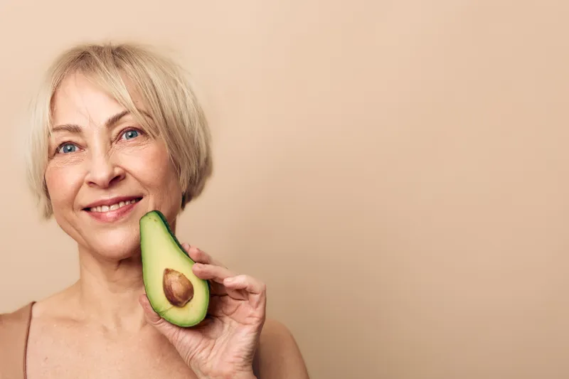 Smiling mature woman with short blonde hair holds a fresh avocado half near her face on beige background. Concept of healthy lifestyle, natural beauty, nutrition, and wellness care.
