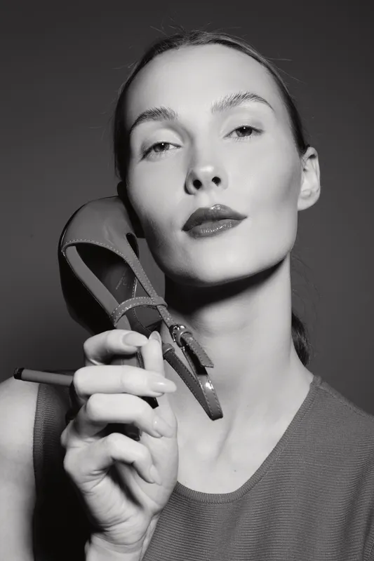 Caucasian woman with pale skin holds high heel shoe near face, posing in studio. Fashion concept with confident model and monochrome lighting.