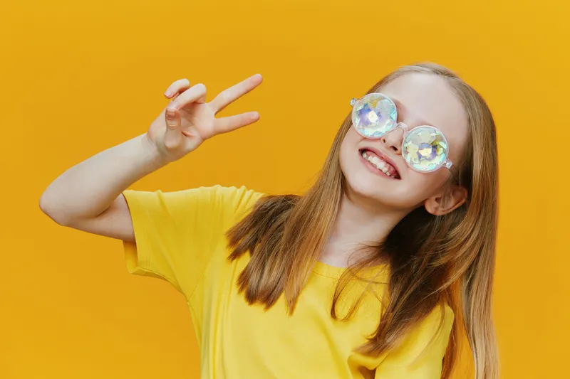 Happy girl in yellow t shirt and colorful sunglasses making peace sign against bright yellow background, showcasing joy and positivity