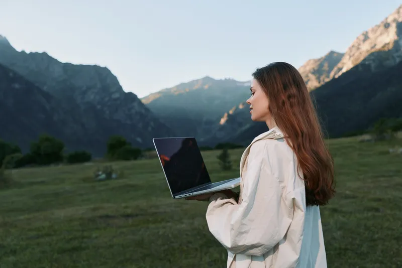 A cheerful professional stands outdoors in a scenic mountain meadow, holding a laptop and smiling as she works, creating a positive, productive, and focused mood.