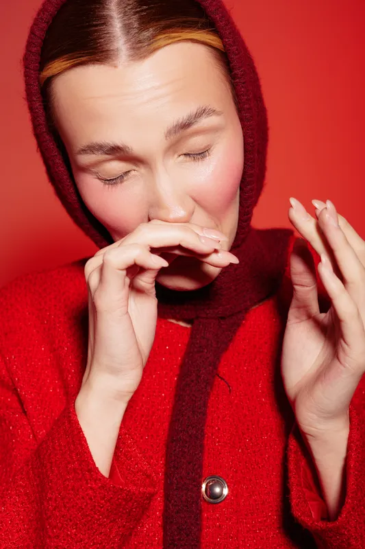 Young woman in red woolen hood and coat sneezes with closed eyes on red background. Concept of cold, allergy, and seasonal illness prevention in winter clothing.