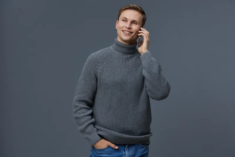 Smiling young man talking on mobile phone while standing in front of plain gray background, casual gray sweater and jeans, relaxed and cheerful demeanor, lifestyle concept, indoor studio shot.