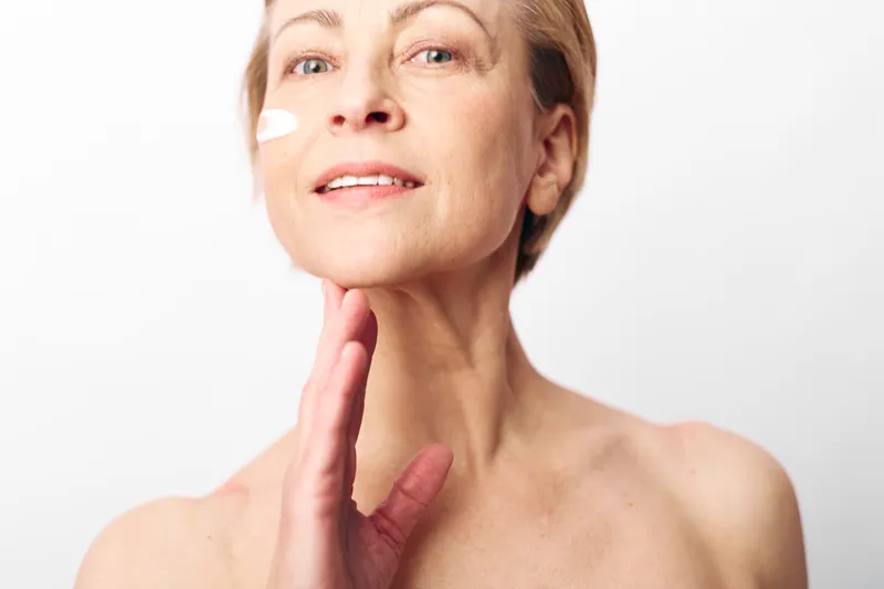 Mature woman with short hair applies moisturizing cream on face, smiling gently. White background and soft natural light highlight skin care and beauty routine for aging skin wellness.
