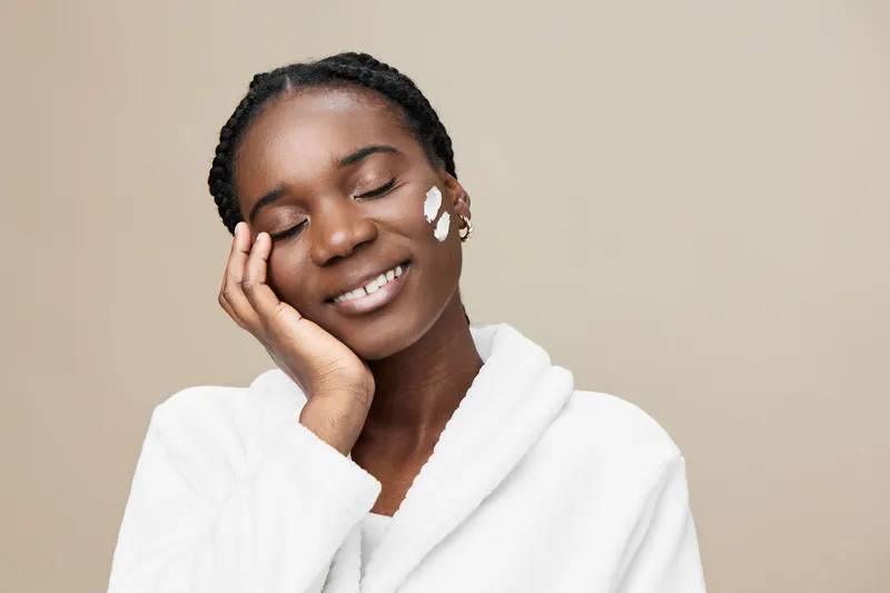 Smiling Black woman applying skincare cream, promoting healthy beauty and self care routines in a calming, neutral toned setting