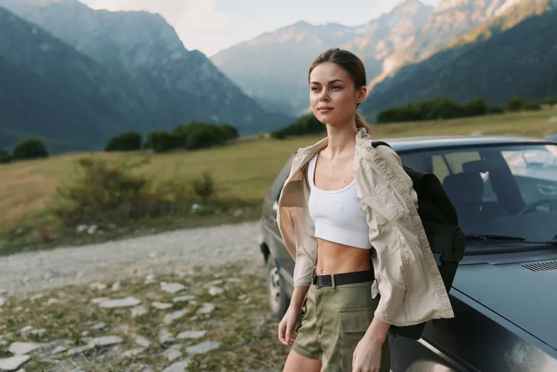Young woman in casual summer clothes stands beside a car on a mountain road enjoying the fresh air and scenic view with a confident and relaxed expression.