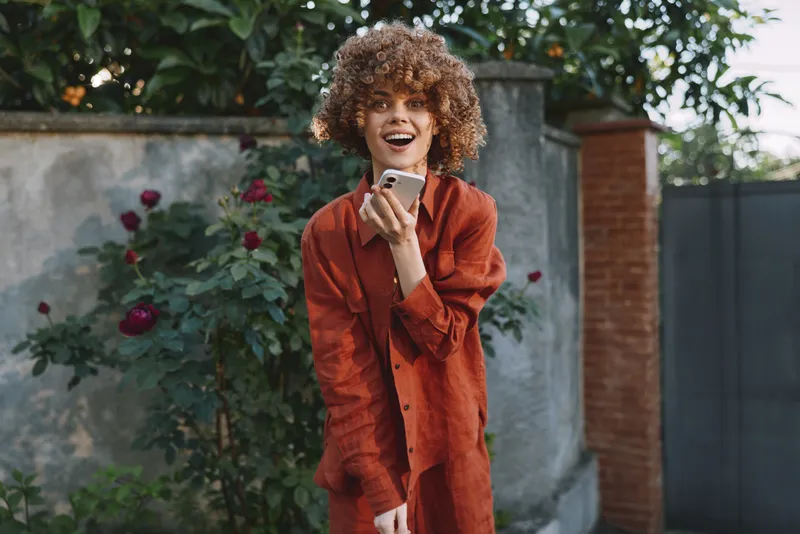 A smiling woman in an orange dress stands outdoors, holding a smartphone. Warm natural light, garden backdrop. Concept for everyday style, happiness, social sharing, and modern life