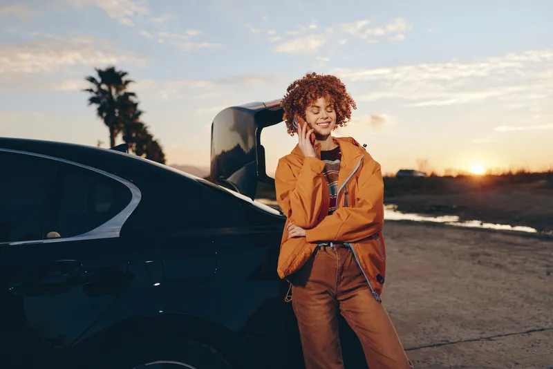 Woman lifestyle near car at sunset, outdoors on road, curly haired woman is smiling, wearing orange jacket, holding small bag, relaxed travel mood, golden hour light warm evening breeze