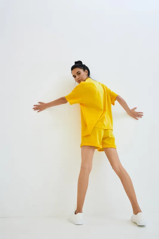 woman in yellow outfit standing against white wall with casual posture and white sneakers, showcasing sporty and relaxed style indoors