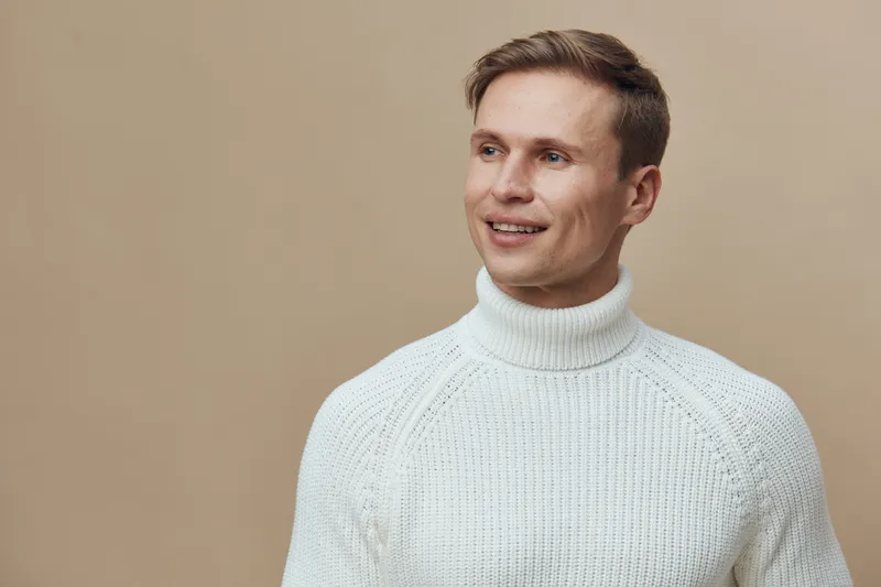 Smiling man wearing white knit sweater looking away isolated on beige background studio portrait. Happiness confidence casual style.