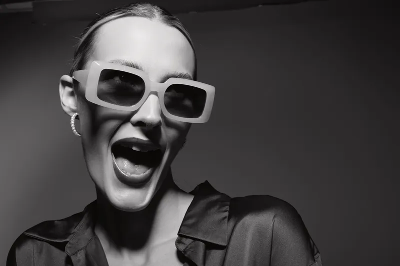 Excited white woman with pale skin wearing large sunglasses and silk blouse smiles widely in studio with dark background.