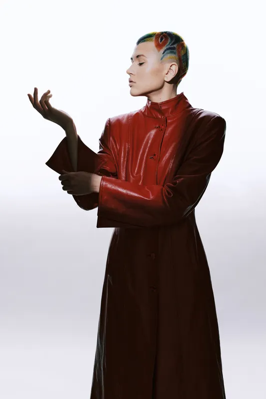 Fashion woman with colorful shaved hairstyle wearing sleek red leather coat posing gracefully against plain white background, modern style and confident attitude.