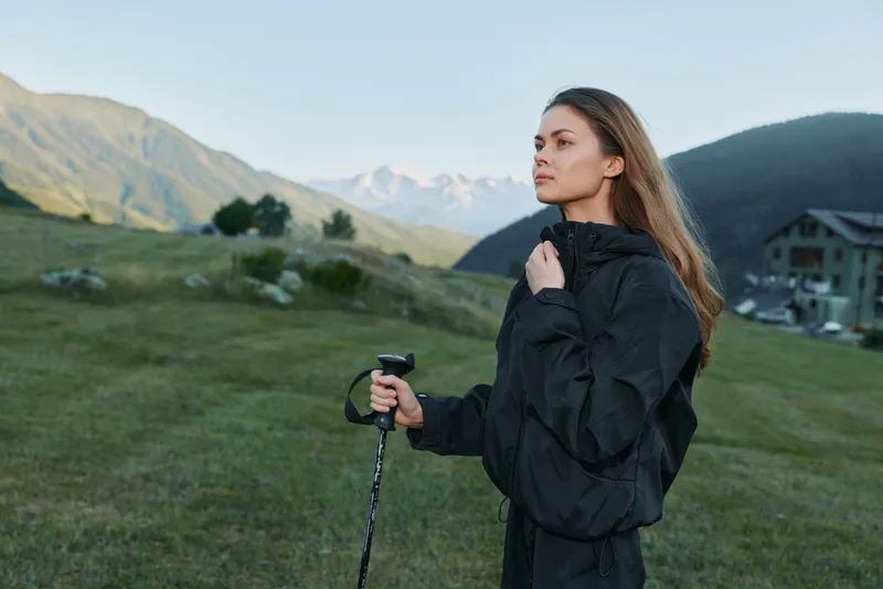 A woman stands outdoors in a mountainous landscape, dressed in a dark jacket. She holds a trekking pole and gazes into the distance with calm resolve, signaling adventure and resilience.