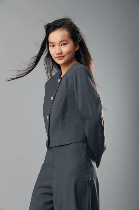 Young woman standing and smiling with long hair blowing, wearing dark gray suit, isolated on gray background. Female model posing in professional outfit with confident expression.
